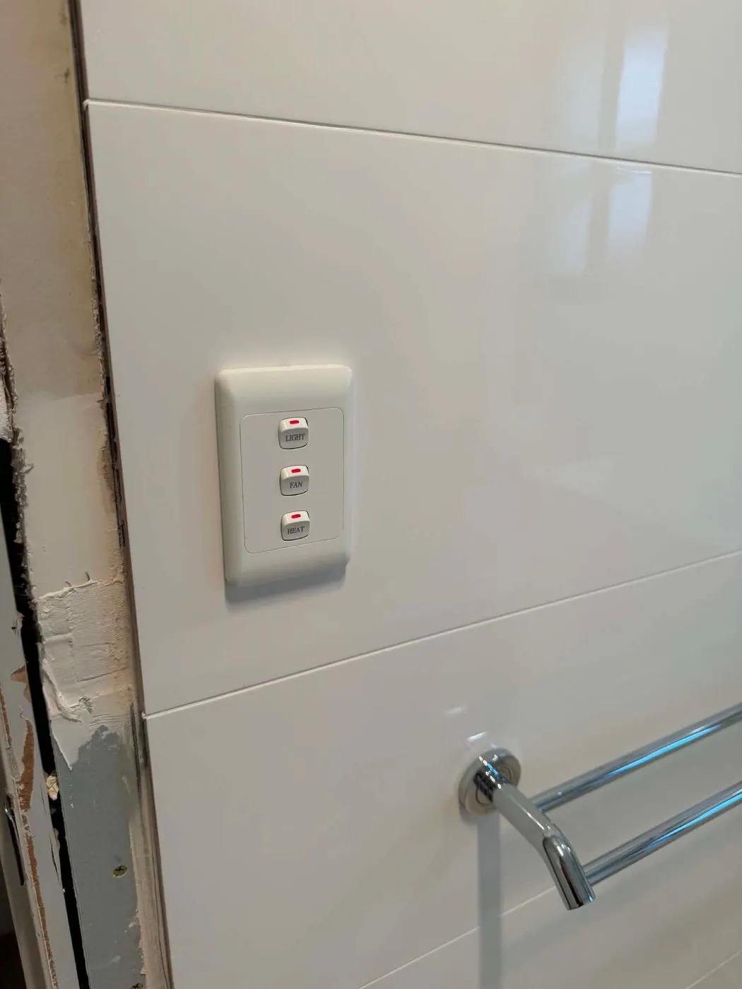 Three-gang switch plate labelled Light, Fan and Heat on a tiled bathroom wall in Wynn Vale
