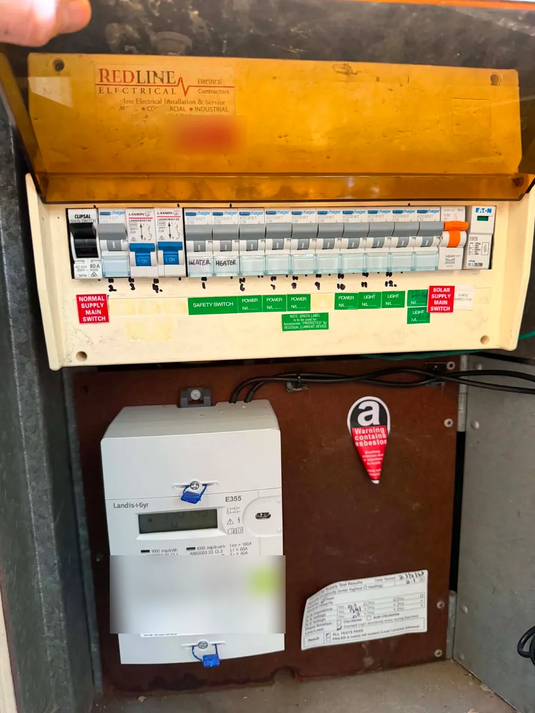 Modern switchboard with breakers, RCD and surge protection above a brown asbestos backing panel carrying a warning sticker