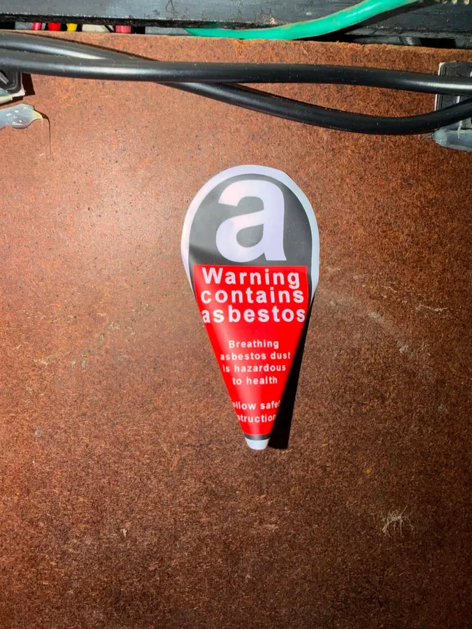 Warning contains asbestos sticker applied to the brown fibrous backing panel inside a switchboard